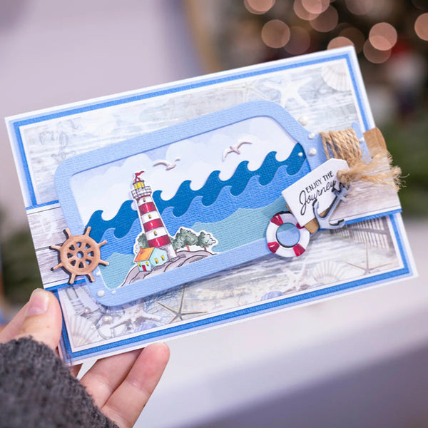 A handmade layered greeting card with a nautical theme, held by a hand. The card features a blue and white striped lighthouse, waves, a ship's wheel, and a small tag that reads 'Gives the Grabs Best'.
