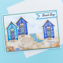 A square greeting card featuring a beach scene with three blue and turquoise sun lounges, a striped beach chair, seashells, and the words 'Beach Days' in a banner, photographed on a light blue background with white envelopes behind it.