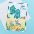 A handmade beach-themed greeting card featuring two blue and green wooden beach huts, two striped deck chairs, and seashells on a yellow sand background, with the text 'Beach Daze!' in the top right. The card is displayed on top of a white envelope against a light blue background.