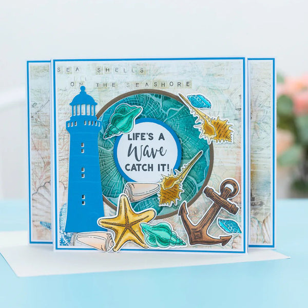 A handmade layered greeting card with a nautical theme, featuring a blue lighthouse, starfish, anchor, seashells, and a teal circular wave design with the text 'LIFE'S A Wave CATCH IT!'.