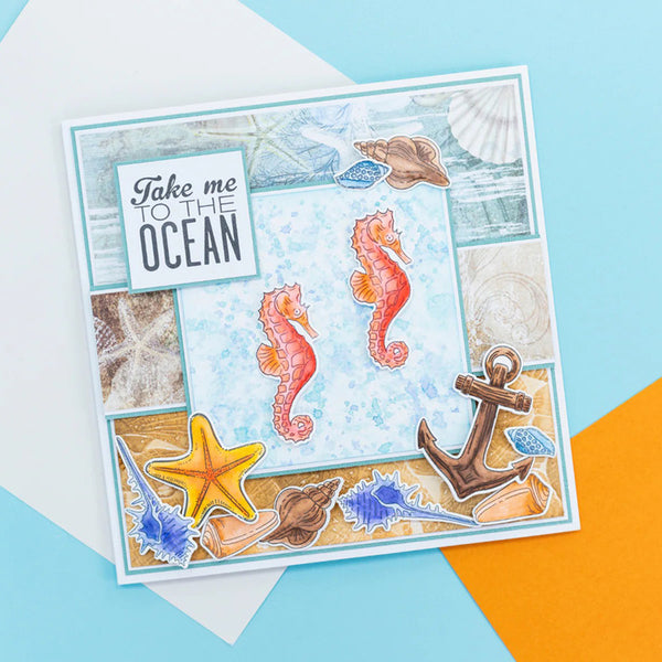 A square handmade greeting card featuring an ocean-themed collage with seahorses, starfish, shells, an anchor, and the phrase 'Take me to the OCEAN' on a light blue background, styled on a flat-lay surface with pastel blue, white, and orange paper accents.
