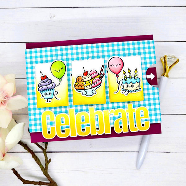 A handmade birthday card with a blue and white gingham background, featuring three cartoon dessert illustrations (a cupcake with a green balloon, a sunda, and a birthday cake) and the word 'Celebrate' in large yellow letters. The card is displayed on a white wood background with decorative flowers and a pen.