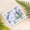 A handmade greeting card with a blue and white plaid background, featuring a central lavender label that reads 'A little magic for you', decorated with blue flowers, green leaves, and butterflies. The card is styled on a light wood surface with green fern leaves.