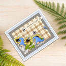 A square greeting card featuring a whimsical garden scene with blue gnomes, sunflowers, butterflies, and the text 'A SEASON OF QUIET JOY', photographed on a light wood surface with green fern leaves around it.
