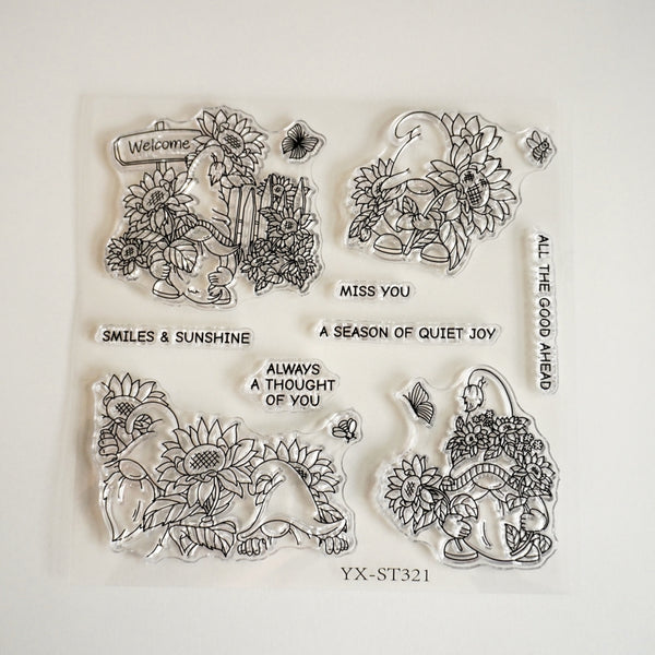 A set of four clear acrylic or rubber stamp designs arranged on a white backing sheet. The stamps feature black line art illustrations of sunflowers, butterflies, and a woman carrying a basket of flowers, with decorative text including 'Welcome', 'SMILES & SUNSHINE', 'A SEASON OF QUIET JOY', 'ALWAYS A THOUGHT OF YOU', and 'MISS YOU'.