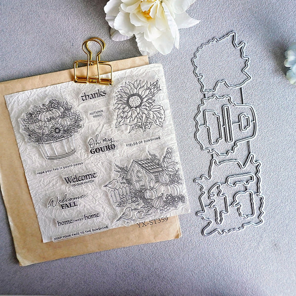 A clear stamp sheet with autumn-themed embossed designs and phrases, clipped to a kraft paper envelope, alongside matching metal stamp dies on a gray surface with a white flower prop.