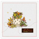 An autumn-themed craft card or sticker sheet featuring a central illustration of a cream-colored cottage nestled among colorful fall leaves, acorns, berries, and a lemon slice. The design is set on a light gray textured background with a stitched border and a brown label reading 'Autumn cottage wishes'.