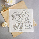 Four metal cookie cutters in irregular floral or abstract shapes arranged on a white square sheet, placed on top of a tan paper envelope with decorative paper flowers and a gold binder clip on a gray surface.