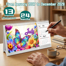A spiral-bound desk calendar displayed on a wooden table, featuring a colorful butterfly and floral cover with the word 'July' and a visible July 2028 page on the right. A person's hands are shown writing in the calendar with a black pen.