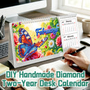 DIY Handmade Diamond Two-Year Desk Calendar shown on a desk with a person's hand writing in an open notebook. The calendar features a colorful diamond-etched design of a blue and orange butterfly on a background of pink roses and green leaves, with 'February' written in blue script.