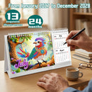A spiral-bound desk calendar displayed on a wooden table, featuring a colorful diamond-pixel bird illustration on the front cover. A hand holds a black pen next to the calendar, with a coffee cup, books, and glasses on the table.