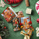 A styled flat-lay of Christmas gingerbread-themed greeting cards and gift boxes on a green textured surface, surrounded by holiday decorations including pine branches, ornaments, cinnamon sticks, and a red ribbon.