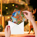A person's hands holding an open white envelope with a colorful Christmas card featuring gingerbread characters, set against a festive blurred background with a tree and a deer figurine.