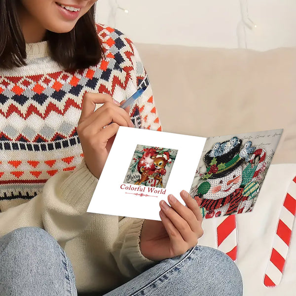 A person sitting on a couch holds a diamond painting canvas featuring a colorful Christmas ornament, with another completed snowman canvas beside it. The person wears a patterned knit sweater and blue jeans in a cozy indoor setting.