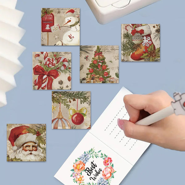 A hand is writing in a floral 'Best Wishes' card, surrounded by six small square Christmas-themed cards or prints with festive illustrations like Santa, snowman, candy canes, ornaments, and a tree. The scene is styled on a light blue background with partial props visible at the edges.