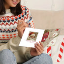 A person in a colorful knit sweater holds a white Christmas-themed card or print featuring a Santa portrait and the words 'Colorful World,' with another patterned card beside it on a light couch.