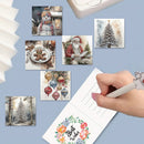A hand holding a white pen writes in an open greeting card, surrounded by six small Christmas-themed art prints on a light blue background. The card features floral 'Best Wishes' writing at the bottom.