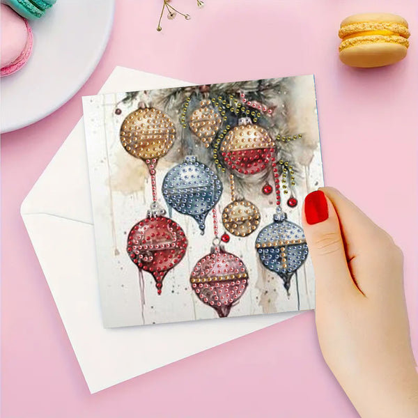 A hand with red nail polish holds a square greeting card featuring a watercolor design of hanging Christmas ornaments, with a white envelope behind it on a pink background.