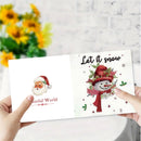 A person holds open a white greeting card or booklet showing two festive holiday designs: a Santa face on the left with the text 'Colorful World,' and a snowman illustration on the right with the text 'Let it snow.' The background is a softly blurred indoor setting with white brick wall and yellow flowers.