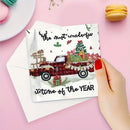 A hand with red nail polish holds a white Christmas greeting card featuring a red pickup truck loaded with holiday decorations and the text 'the most wonderful time of the YEAR'. A white envelope lies behind the card on a pink background, with macarons and small decorative items in the corners.