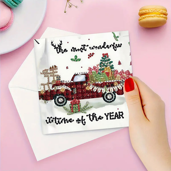 A hand with red nail polish holds a white Christmas greeting card featuring a red pickup truck loaded with holiday decorations and the text 'the most wonderful time of the YEAR'. A white envelope lies behind the card on a pink background, with macarons and small decorative items in the corners.