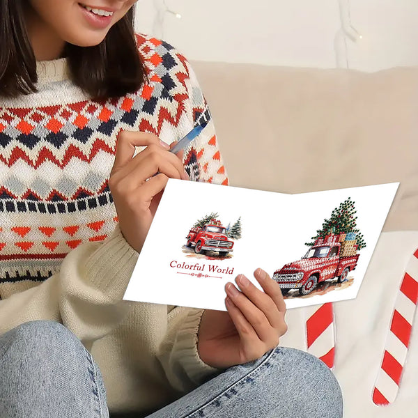 A person in a patterned knit sweater holds a white greeting card featuring two red vintage trucks carrying Christmas trees, with the text 'Colorful World' on the card.