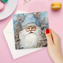 A hand with red nail polish holds a square card featuring a festive illustration of a bearded figure in a blue hat, overlaid with white diamond painting rhinestones, on a pink background with a white envelope and macarons.