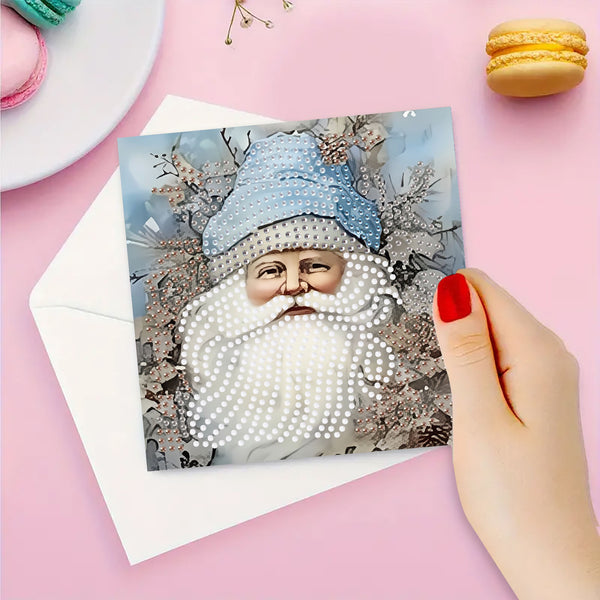 A hand with red nail polish holds a square card featuring a festive illustration of a bearded figure in a blue hat, overlaid with white diamond painting rhinestones, on a pink background with a white envelope and macarons.