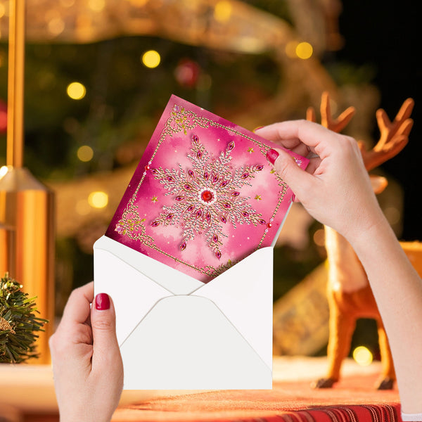 A person holds an open white envelope and a pink holiday card with a jeweled snowflake design, with a blurred Christmas tree and decorative reindeer in the background.