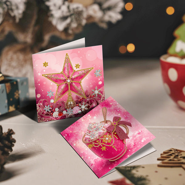 Two pink Christmas greeting cards displayed on a tabletop with holiday decor in the background. One card shows a large glittery gold star, and the other shows a pink gift bag with a bow and gold stars.