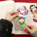 A person holds a Christmas stocking-shaped diamond painting or sticker with Santa and a snowman design over an open book. The stocking has green, pink, white, and red details with rhinestone-like embellishments.