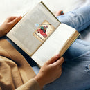 A person lying on a bed or sofa holding an open book, with a small square Christmas-themed card or bookmark tucked into one page. The card shows a red vintage truck carrying a green Christmas tree and the words 'Merry Christmas!' in black script.