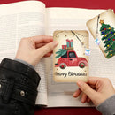 A person holds a small rectangular Christmas card featuring a red pickup truck carrying wrapped presents, with the text 'Merry Christmas' at the bottom. An open book lies underneath, and a second card with a hanging Christmas tree design is visible to the right.