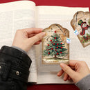 A person holds two vintage-style Christmas paper bookmarks or tags over an open book. One tag features an illustrated green Christmas tree with red ornaments, and the other shows a snowman on a decorated tree.