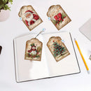 Three Christmas-themed cross-stitch embroidery patches displayed on a white desk, with one patch shown inside an open notebook and two others placed above it. The patches feature festive designs including Santa Claus, a red gift box, a snowman, and a Christmas tree on a parchment-style background.