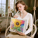 A woman seated in a rope swing holds a clear tote bag decorated with a colorful floral artwork featuring a large yellow sunflower and pastel rainbow background.