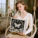 A woman seated in a rope swing holds a black tote bag decorated with a large butterfly and floral motifs in white, silver, and gold rhinestone-like embellishment. The setting is a softly lit indoor space with plants and warm natural light.