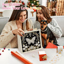 A woman holds a decorative gift bag featuring a black background with a large silver butterfly, white flowers, and hanging pearl-like embellishments. Another woman sits beside her holding a small black-and-tan dog, with wrapped presents and holiday decorations on a table in a cozy indoor setting.