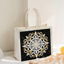 A white fabric tote bag with a large black front panel featuring an ornate white and gold mandala-style embellished design. The bag has white handles and is photographed in a softly lit indoor setting with neutral decor.