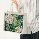 A person wearing a white and gray striped shirt holds a clear plastic tote bag with a green tropical leaf print embellished with rhinestones and white pearl-like bead details.