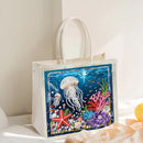 A white canvas tote bag with cream-colored handles, featuring a colorful, rhinestone-embellished underwater scene on the front with a jellyfish, starfish, coral, and sea life, set against a warm, sunlit background.