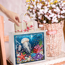 A clear plastic gift bag with a white handle and a square printed underwater scene featuring a jellyfish, coral, and starfish, being held by a hand next to a woven basket of flowers.