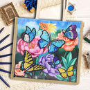 A square canvas or glass panel artwork featuring colorful painted butterflies over pink, purple, yellow, and orange flowers, styled on a white surface with decorative props including blue pencils, a small framed print, a wrapped gift, and jewelry accessories.