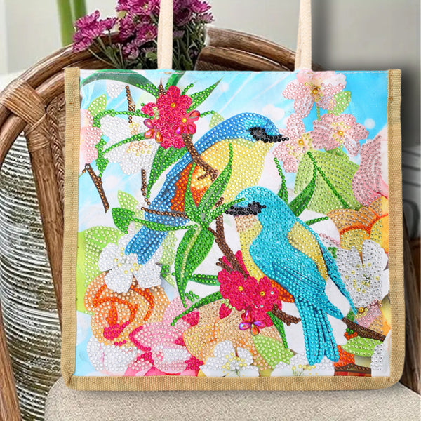 A canvas tote bag with a colorful diamond painting design of two bluebirds, pink flowers, and green leaves, displayed on a wicker chair.