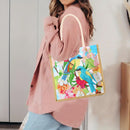 A woman wearing a dusty pink cardigan and light blue jeans carries a square canvas tote bag with colorful embroidered birds and flowers. The bag has a light beige handle and trim, set against a neutral indoor background.
