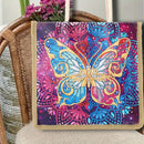 A canvas tote bag with a vibrant, colorful butterfly design and intricate mandala patterns, resting on a wicker chair.