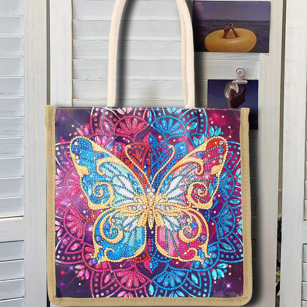 A canvas tote bag with cream-colored shoulder straps, featuring a large, vibrant, rhinestone-embellished butterfly design on a purple and pink mandala background. The bag is displayed hanging against a white wooden shutter door with a small picture frame clipped to the side.
