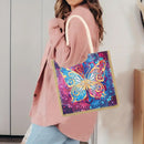 A woman wearing a pink cardigan and light blue jeans carries a square canvas tote bag with a colorful butterfly design on a purple background. The bag has cream-colored handles and a gold-colored stitched border.