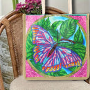 A square canvas art piece featuring a colorful beaded butterfly on green leaves, displayed on a woven rattan chair with flowers in the background.