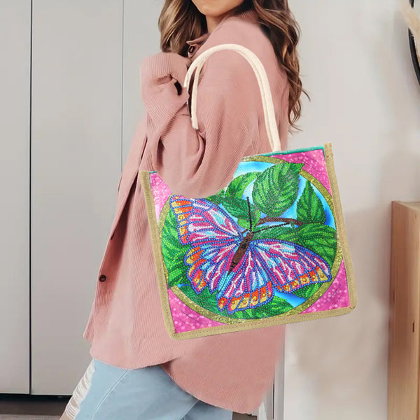 A woman wearing a pink cardigan and light blue jeans carries a colorful tote bag with a glittery butterfly design on a pink background. The bag has cream-colored shoulder straps and is shown in an indoor setting.